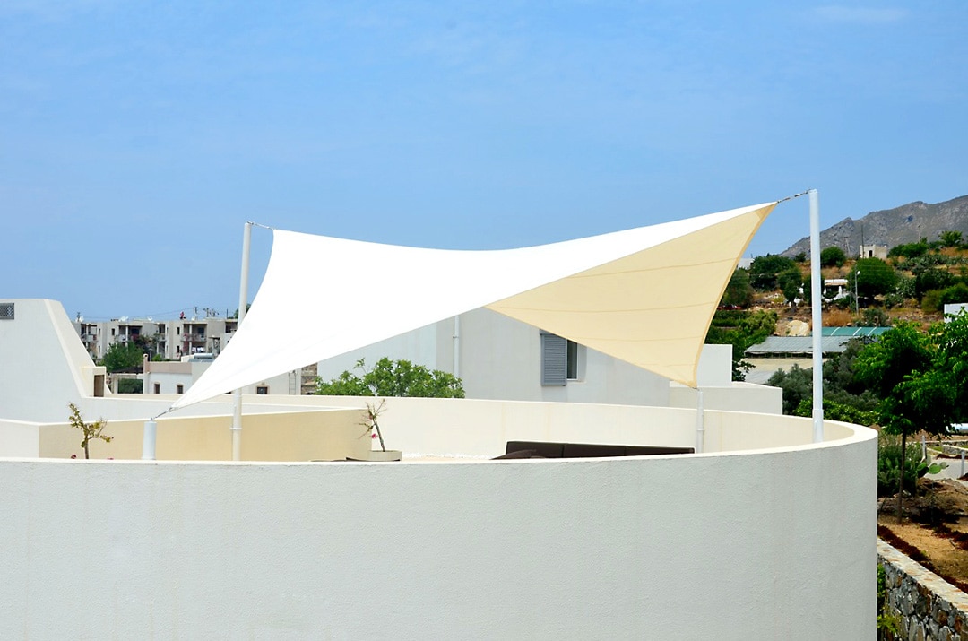 A large, elegantly designed shade sail provides shelter over a rooftop terrace. The structure is anchored by sleek poles, casting gentle shadows on the smooth white surface below. The background features a landscape of white buildings and greenery under a clear blue sky.