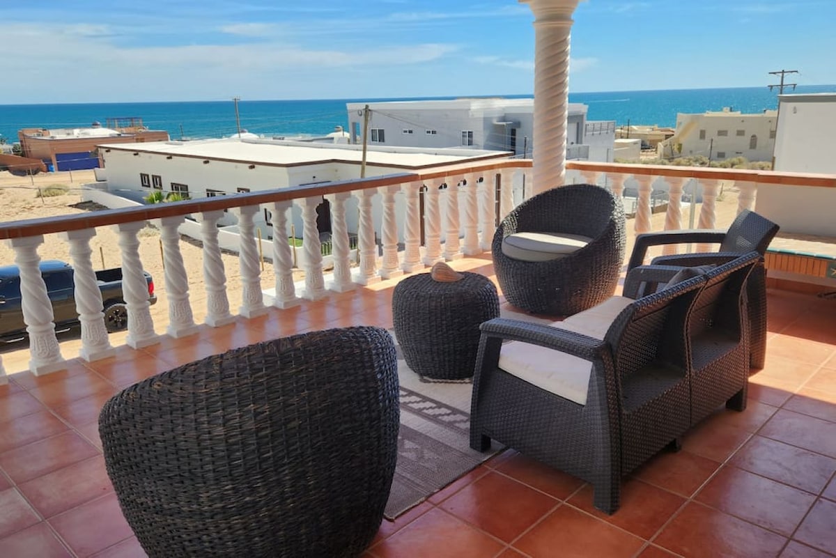A balcony seating area features two wicker chairs and a small table, offering a relaxing view of the Sea of Cortez. The tiled floor reflects natural light, while a railing provides safety. Ocean waves and distant buildings are visible in the background.