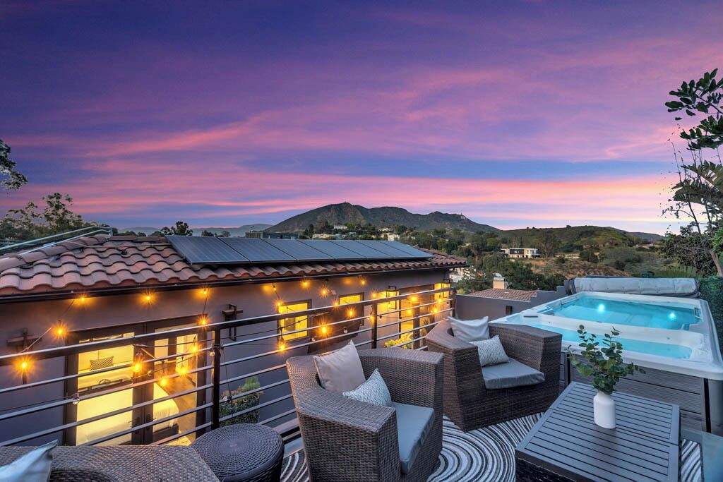 A spacious outdoor terrace features cozy seating arrangements with plush cushions and a hot tub, surrounded by decorative string lights. The backdrop consists of rolling hills and the Hollywood sign against a vibrant sunset sky, creating a calm and inviting atmosphere.
