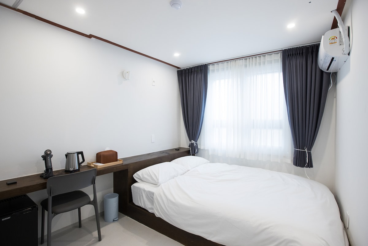 A cozy guest room features a spacious bed dressed in white linens. Natural light filters through sheer curtains, enhancing the serene atmosphere. A small desk is positioned beside a refrigerator and a kettle, while a chair provides a comfortable spot for relaxation.