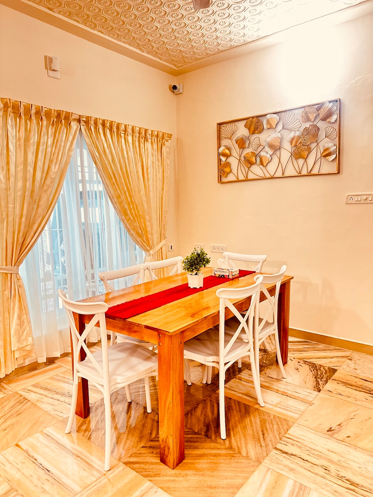 A bright dining area features a wooden table surrounded by four white chairs. Natural light fills the space, highlighting a decorative wall art piece above the table. Curtains frame a window that offers a view of the outside.