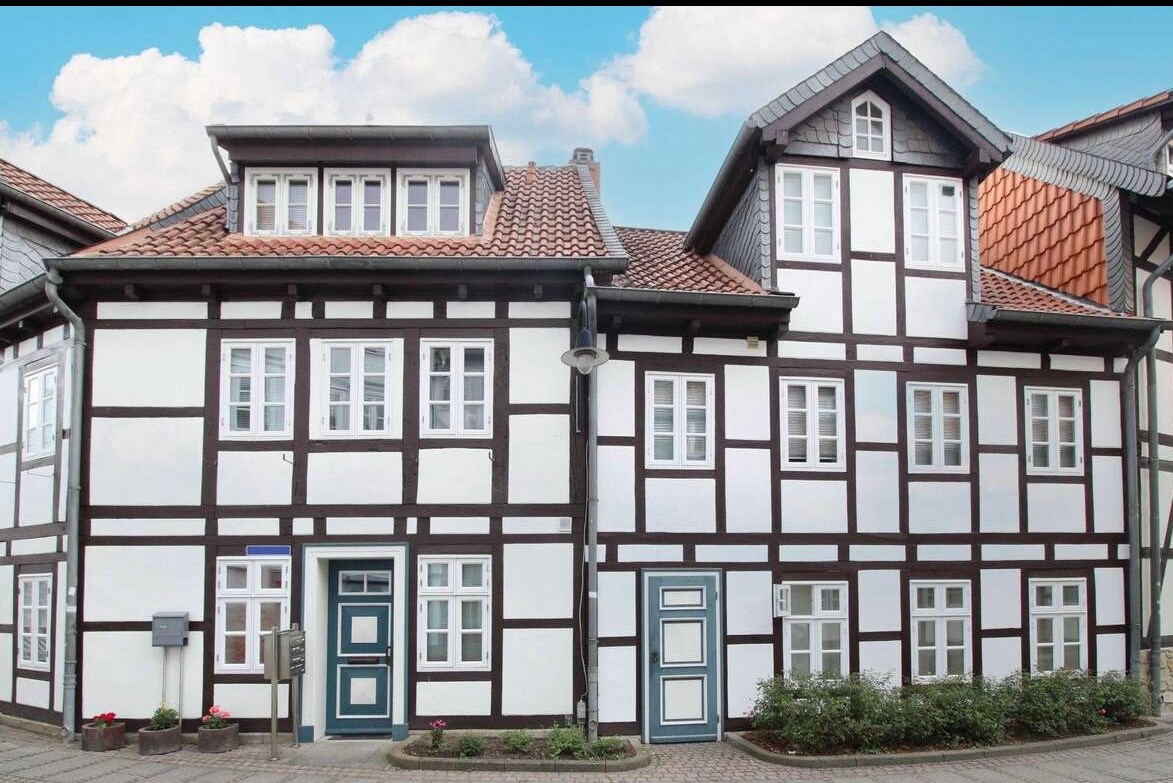 A historic, half-timbered building showcases a charming facade with contrasting dark wood and light-colored walls. Multiple windows are present, each framed in white, while blue doors add a subtle touch of color. A well-maintained garden borders the entrance.