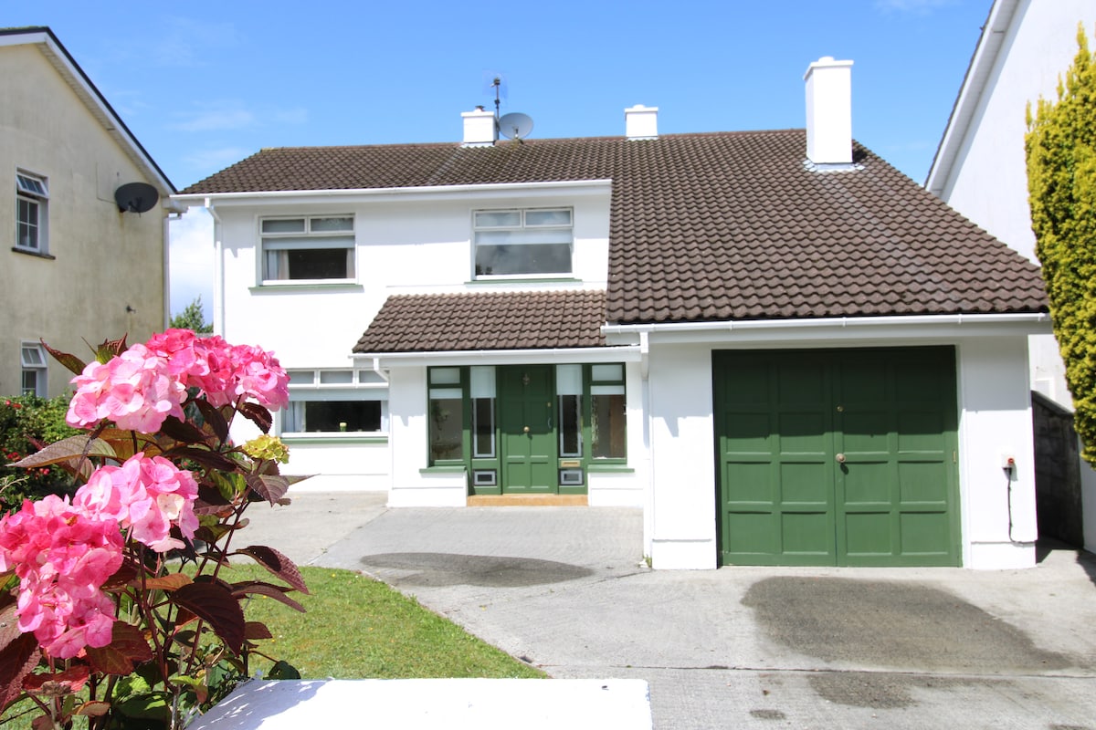 A detached two-story home is set against a clear blue sky. The exterior is painted white with a brown tiled roof. Green front doors are complemented by matching garage doors. Colorful pink flowers are visible in the foreground, adding a touch of nature.