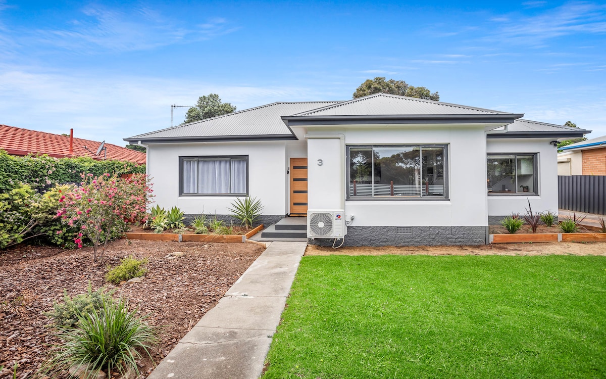 The exterior of a newly renovated single-story home features a well-manicured lawn and landscaped gardens. A paved walkway leads to the entrance, which is highlighted by a modern door beside a large window. An air conditioning unit is visible on the side of the house.