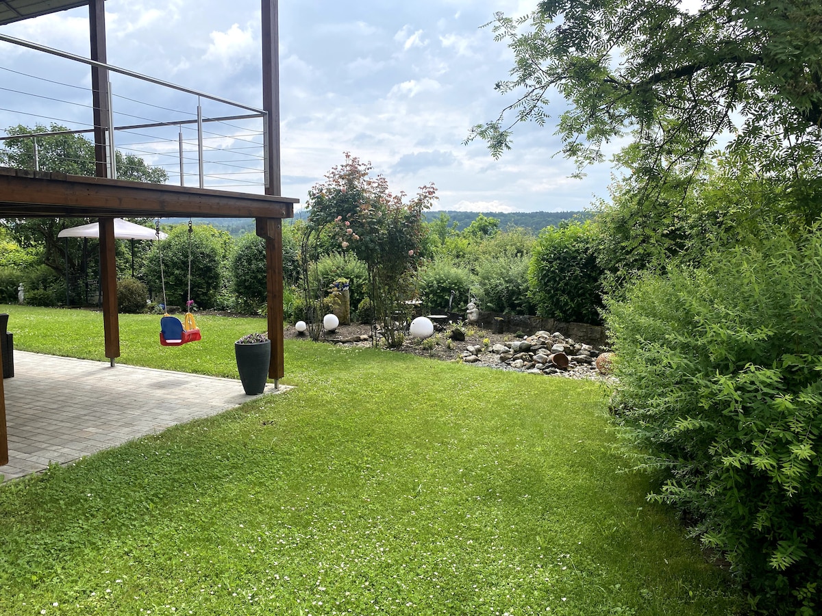 A spacious garden area is visible, featuring green grass and decorative plantings. A wooden structure extends overhead, providing shade. A small swing set is positioned on the left, while ornamental white globes dot the landscape, contributing to the garden's serene ambiance.