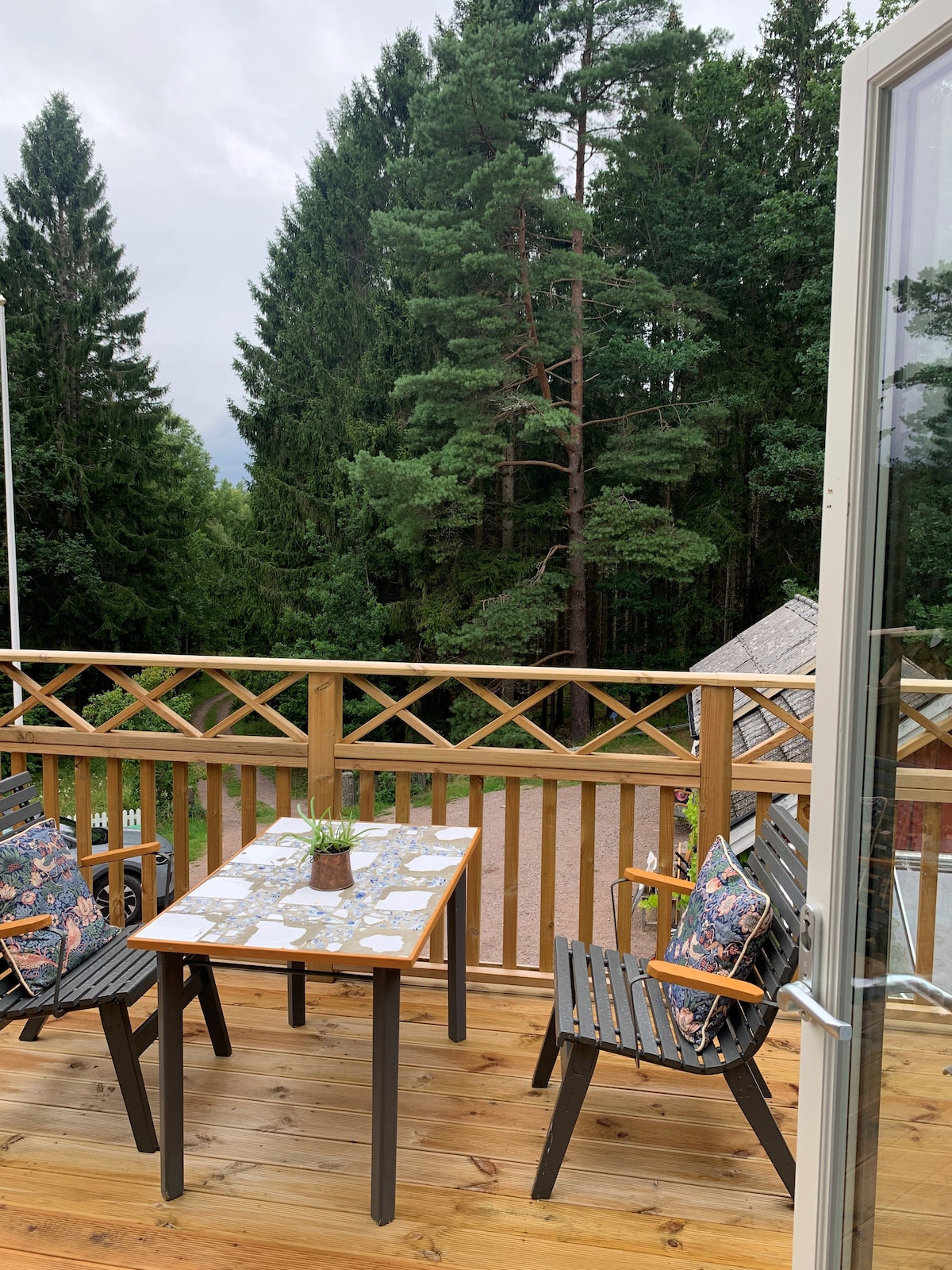 An outdoor patio features a wooden table accompanied by two black chairs, adorned with floral pillows. The railing offers views of tall evergreen trees, creating a natural backdrop. A small potted plant sits on the table, contributing to the inviting outdoor space.