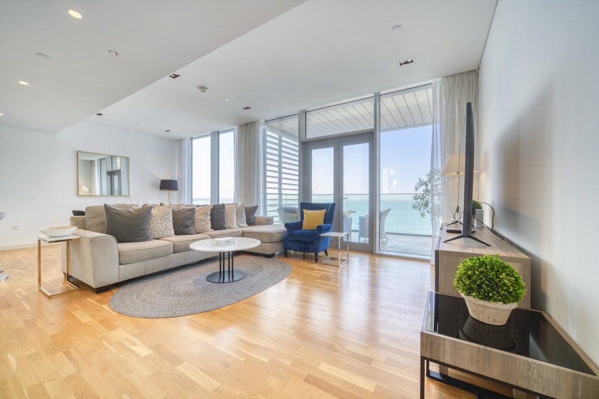 The spacious living area features modern furnishings, including a comfortable sofa, coffee table, and decorative rug. Floor-to-ceiling windows allow for ample natural light and provide a view of the ocean. A sleek entertainment unit is complemented by minimalist decor, enhancing the room's tranquility.