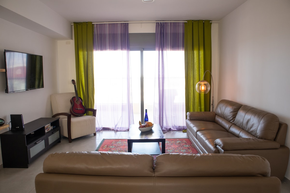 A modern living room features two leather couches facing a coffee table adorned with fruit. A flat-screen TV is mounted on the wall opposite. Sheer curtains in shades of green and purple frame the large window, allowing natural light to fill the space.