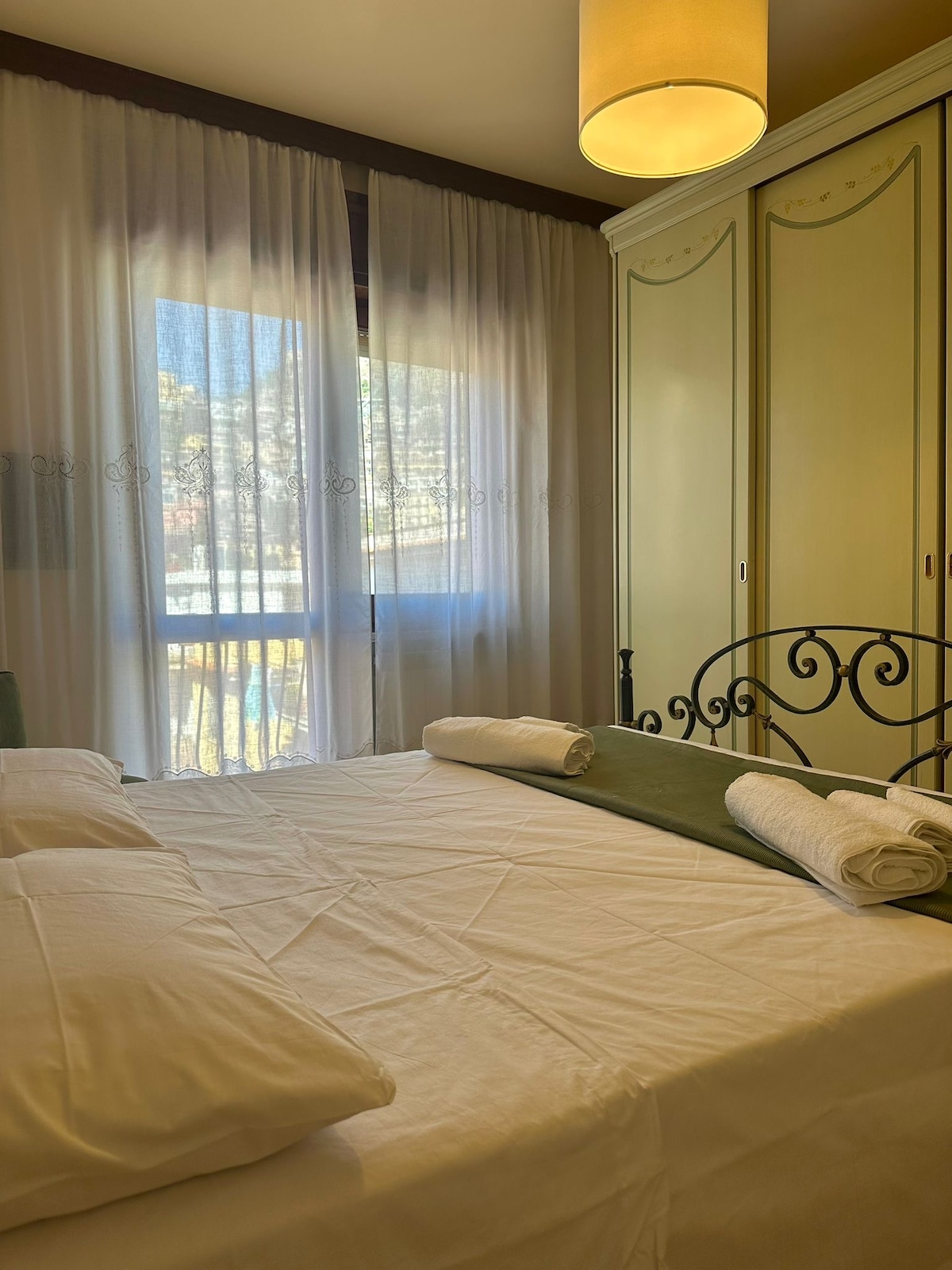 A cozy bedroom features a spacious bed covered with white linens, complemented by neatly rolled towels. Light filters through sheer curtains, illuminating the room. An elegant wardrobe stands against the wall, offering ample storage space, while a soft light fixture hangs from the ceiling.