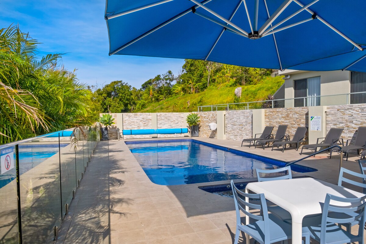 A well-maintained swimming pool area is showcased, featuring clear blue water and surrounded by comfortable sun loungers. A large blue umbrella provides shade over a dining table set for six, enhancing the open and inviting atmosphere. Lush greenery borders the space, complementing the serene environment.