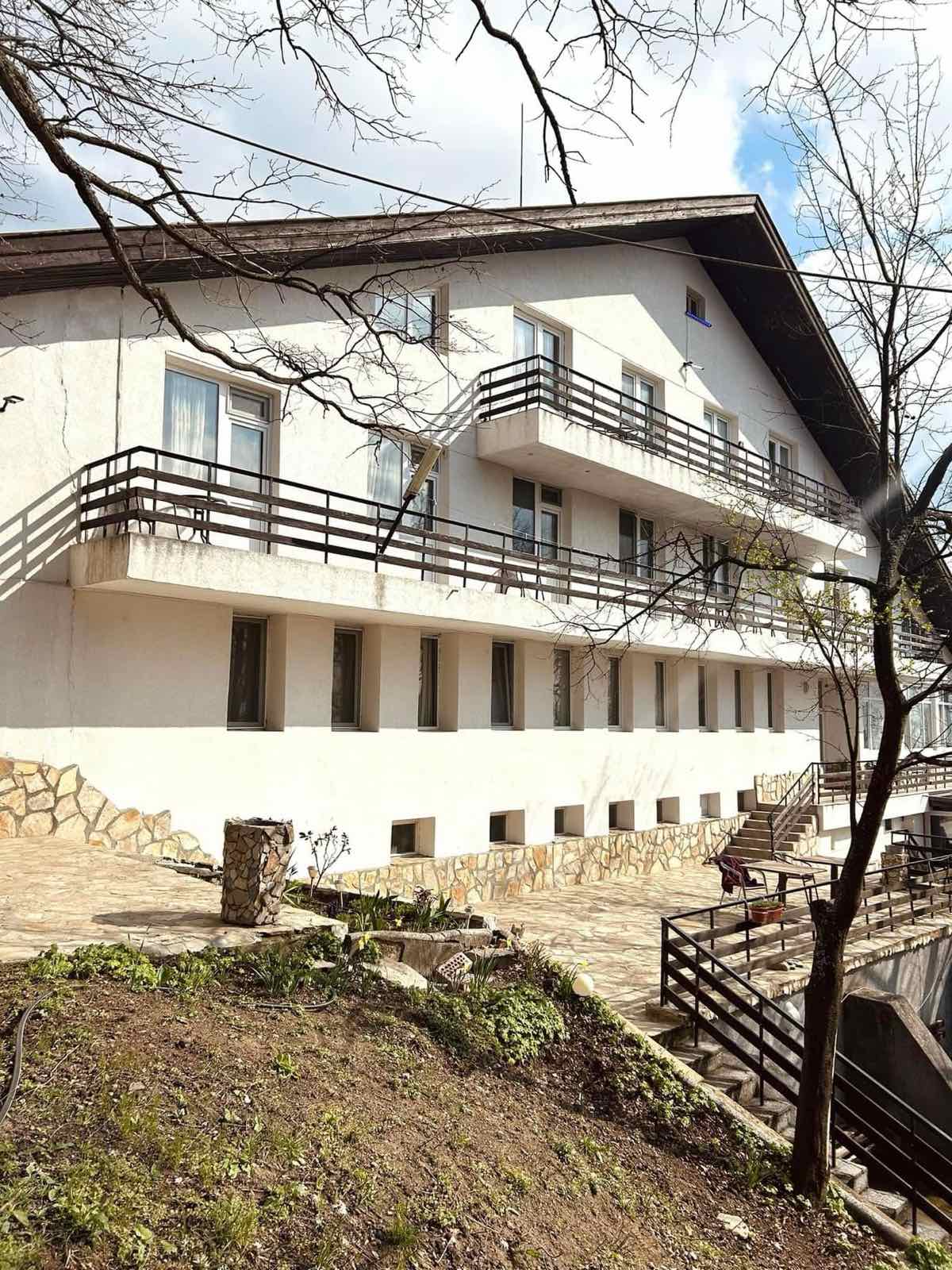 The exterior of a three-story building is showcased, featuring a simple white facade with multiple balconies. A stone pathway and landscaped area are visible in front, complemented by bare trees and a cloudy sky overhead.