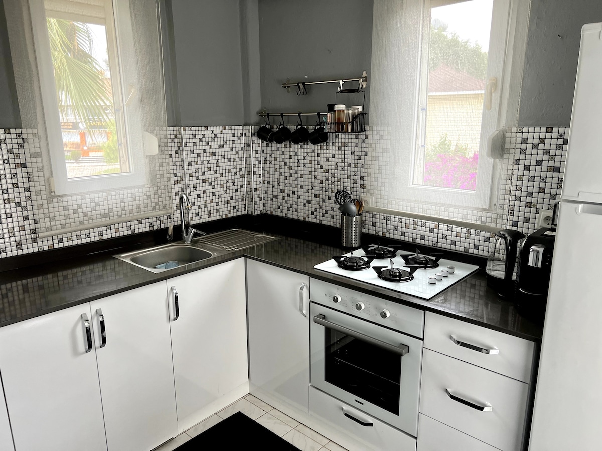A modern kitchen is equipped with a stainless steel stove and an oven. The cabinetry features sleek white finishes, while a mosaic tile backsplash complements the space. Natural light enters through two windows, which overlook a garden. Essential kitchen tools are neatly arranged on a rack.
