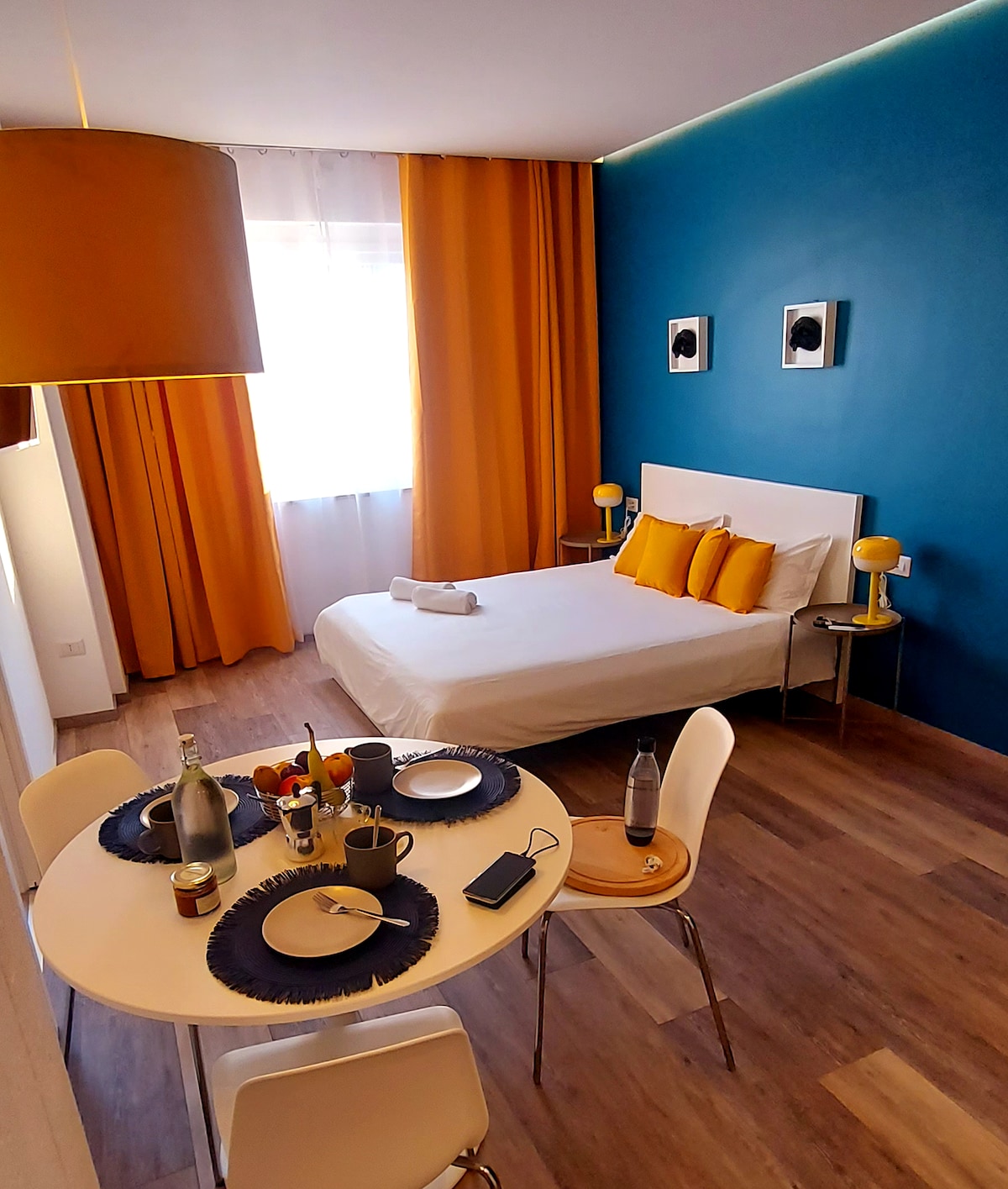 A bright and modern studio features a spacious bed adorned with decorative pillows. A small dining table is set for two, showcasing dishes and beverages. Warm curtains complement a blue accent wall, while a large window provides natural light.