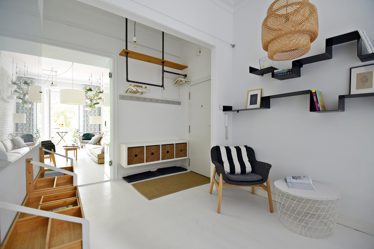 An entryway is shown with bright white walls and a wooden ceiling fixture. A comfortable chair sits beside a round coffee table. Shelves display decorative items, while a storage unit with woven drawers is present. Natural light enters from an adjacent room with greenery.