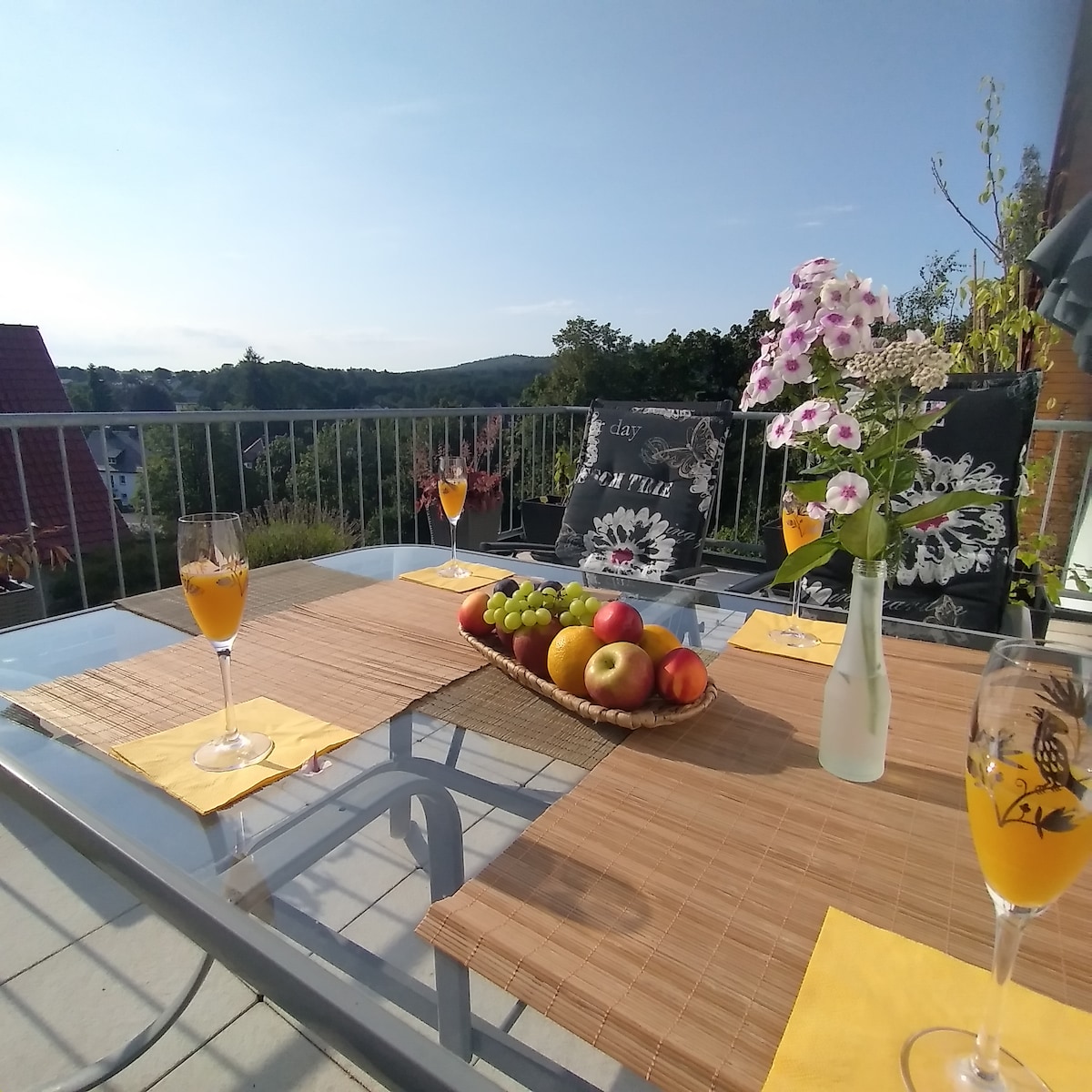 A spacious outdoor dining area features a glass table adorned with drinks and a decorative fruit basket. Colorful flowers are in a vase, enhancing the serene setting with distant greenery and hills visible in the background under a clear blue sky.