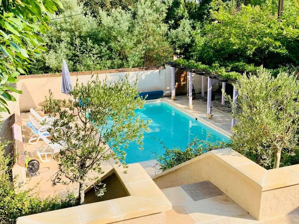 A spacious swimming pool is surrounded by lush greenery, providing a serene atmosphere. Sun loungers and umbrellas are placed around the pool area, which is enhanced by natural shade from nearby trees. Soft stone steps lead to the pool from an elevated vantage point.