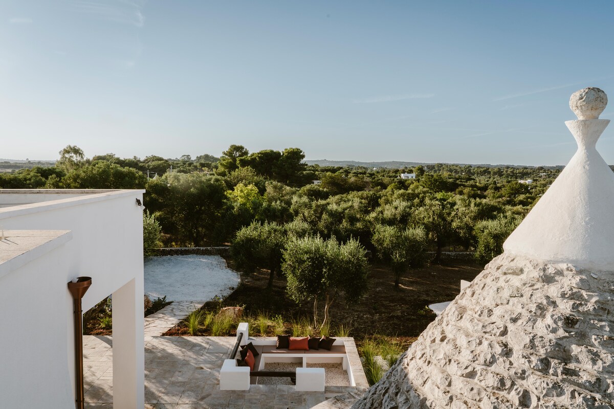 A panoramic view showcases a lush landscape filled with olive trees, extending into the distance. A cozy seating area is visible on a patio, and a traditional conical roof structure is positioned nearby, contributing to the serene outdoor environment.