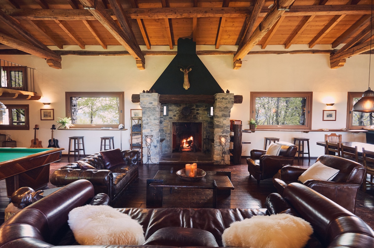 A spacious living area features leather sofas arranged around a rustic coffee table. A stone fireplace serves as a focal point, with wood accents and large windows allowing natural light to illuminate the space. A pool table is visible in the background, enhancing the relaxing atmosphere.