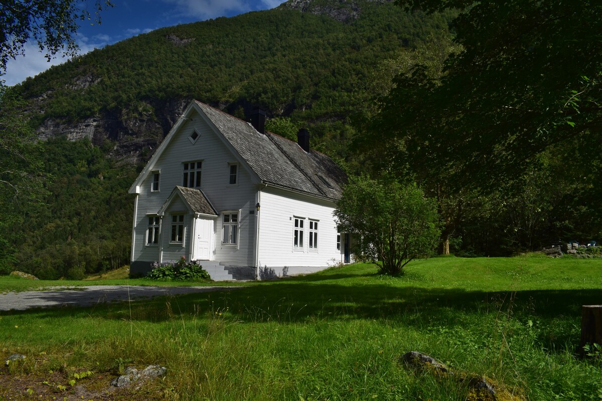 The large white house is set against a backdrop of lush green mountains. Its two-story façade features several large windows, allowing natural light to illuminate the interior. Surrounded by a spacious lawn, the house offers a peaceful atmosphere in a natural setting.