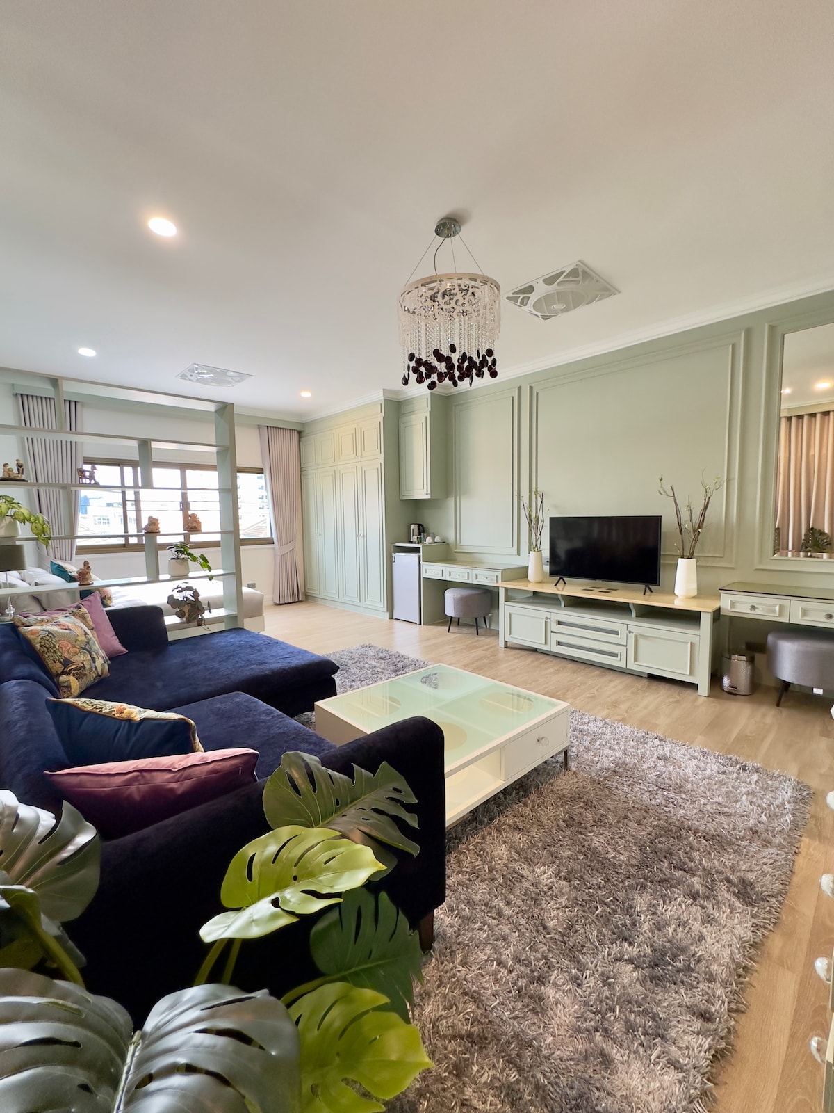 A well-lit living area is adorned with soft blue sofas and a glass coffee table. A smart TV is mounted on the wall opposite the seating. Large windows allow natural light to flood the space, complemented by decorative plants and a stylish chandelier above.