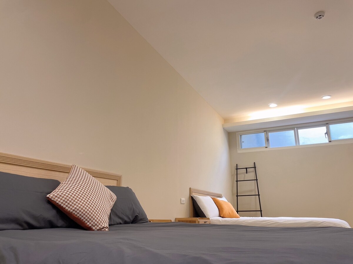 A spacious room features two beds, one with a dark grey blanket and a decorative pillow, and another with crisp white bedding. Natural light enters through multiple windows, enhancing the calming neutral tones of the walls. A tall ladder rests against the wall, adding a unique element.
