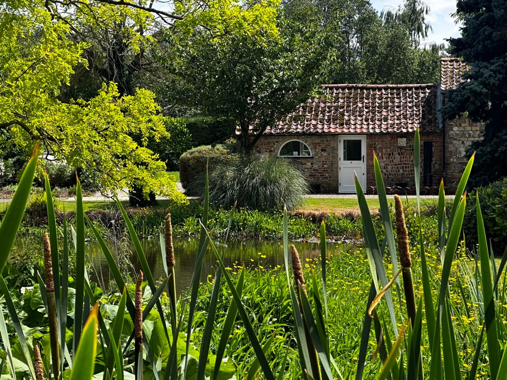 A view of the private annexe is framed by lush greenery and tall plants. The structure features a rustic brick exterior with a tiled roof. A tranquil lake is visible in the foreground, enhancing the serene atmosphere of the setting.