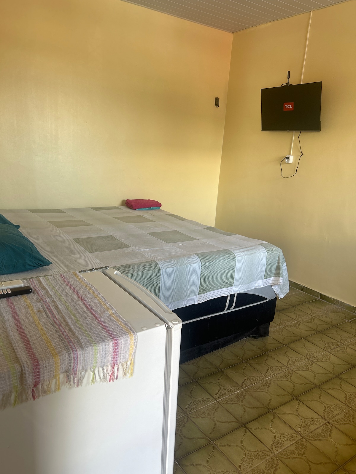 A cozy room features a spacious bed covered with a light-colored blanket. A small television is mounted on the wall. A compact refrigerator is positioned next to the bed, with a colorful towel neatly placed on top. The room is illuminated by natural light.