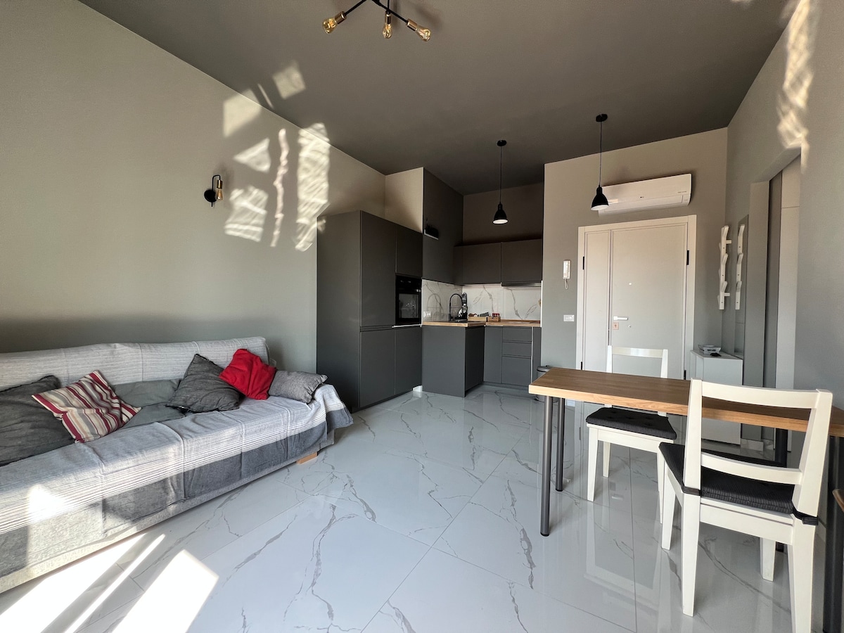 A modern living area features a gray sofa with red cushions and a dining table with two white chairs. A sleek kitchen is visible in the background, complemented by polished floor tiles and soft overhead lighting.
