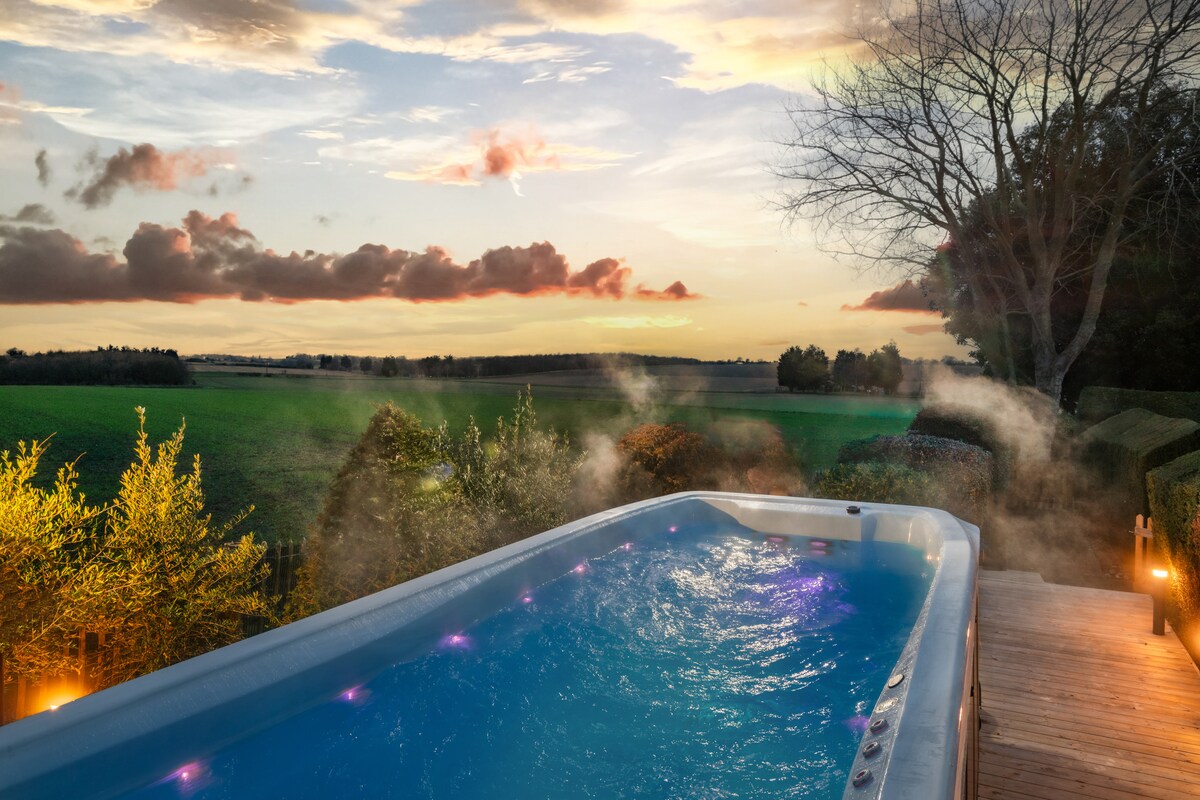 A private spa pool with swim jets is situated outdoors, surrounded by landscaped gardens. Soft steam rises from the water, contrasting with the twilight sky, which features colorful clouds over green fields. Gentle lighting highlights the area, creating a soothing environment.