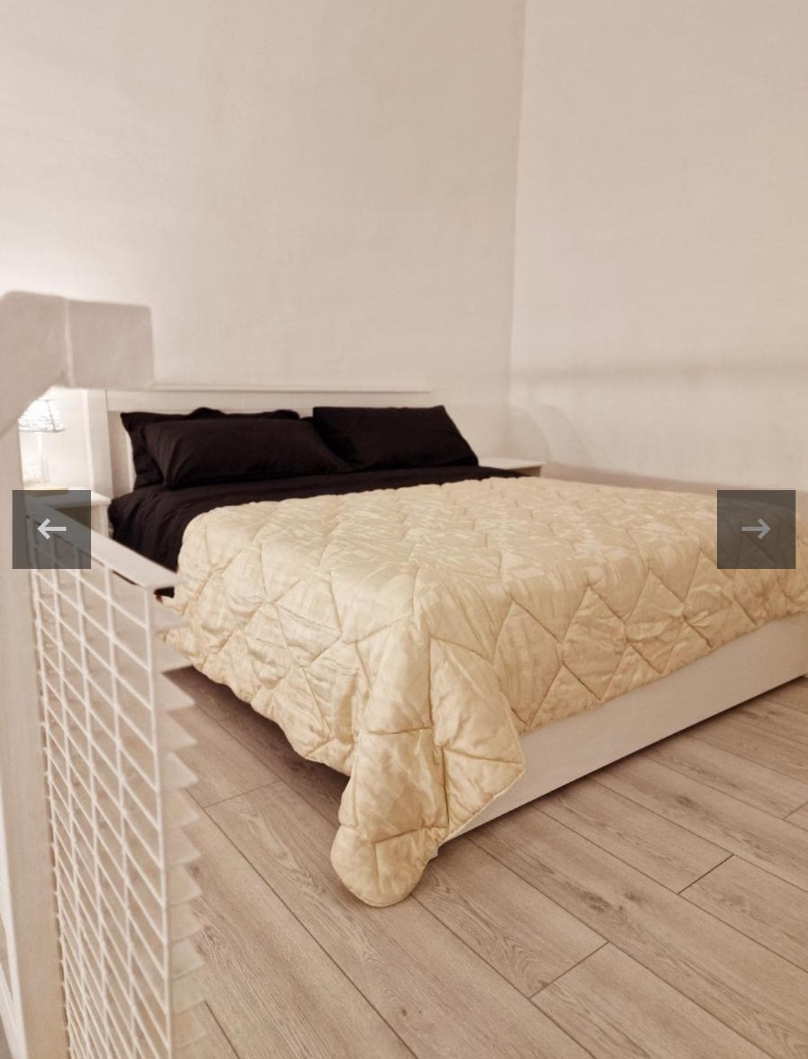 A spacious bed is dressed in a light quilted coverlet, complemented by dark pillowcases. The minimalist design is accented by a light wooden floor and neutral walls, creating a calm and relaxing ambiance. A bedside lamp provides soft illumination.