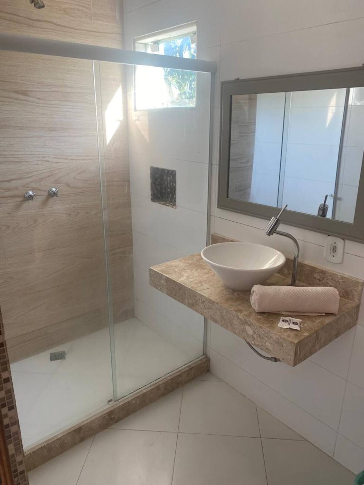 A modern bathroom is featured with a spacious glass shower enclosure. A sleek wall-mounted sink with a bowl design sits atop a light-colored countertop. Natural light enters through a small window, and a mirror is positioned above the sink.
