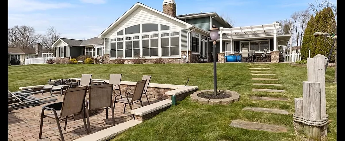 A spacious outdoor area features a well-kept lawn and a stone fire pit surrounded by several seating chairs. A pathway leads to the house, which showcases large windows and a stone foundation, providing ample natural light to the interior.