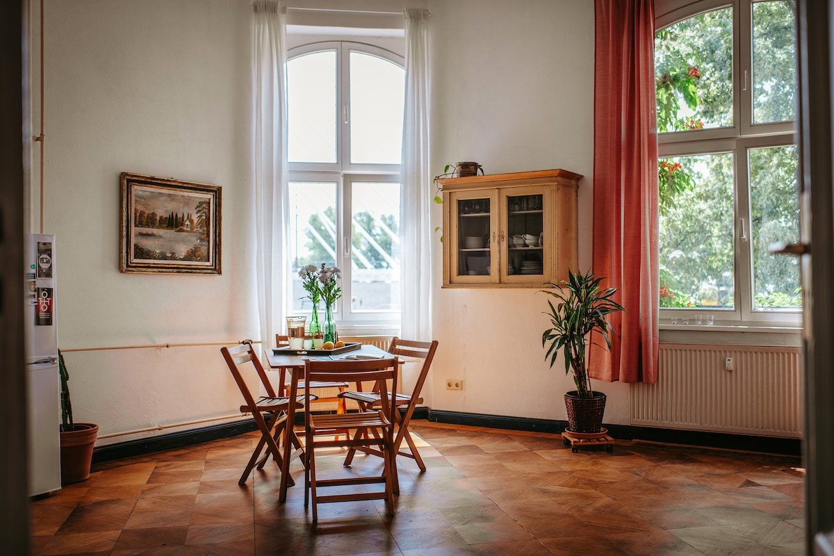 A dining area features a wooden table surrounded by four folding chairs. Large windows allow natural light to fill the space, framed by light curtains. A small plant and a cabinet with glass doors add a touch of greenery and organization to the room.