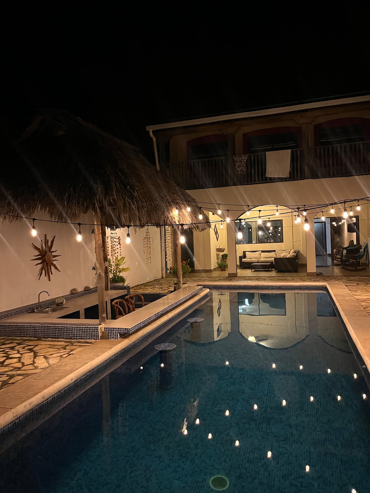 The pool area is illuminated by string lights, casting a warm glow over the water. A thatched roof structure provides shelter near the pool, while comfortable seating is arranged nearby. The architecture of the house is visible in the background, creating a welcoming evening ambiance.