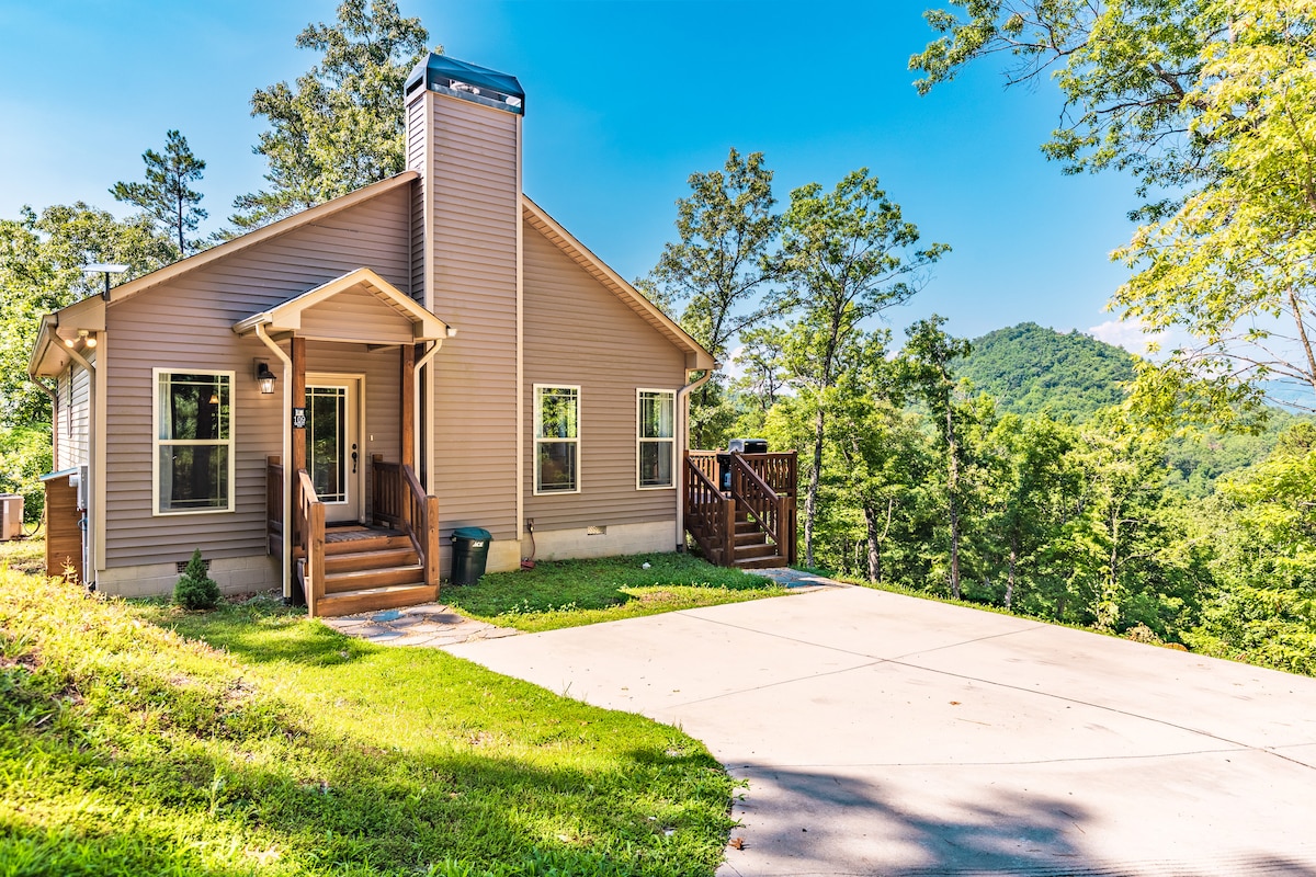 A charming single-story house is surrounded by lush greenery, showcasing a well-maintained driveway leading to the front entrance. Large windows allow natural light into the inviting interior, while a gentle slope of the hillside offers a serene backdrop.