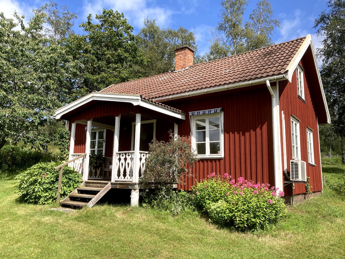 A charming red cottage is set in a lush green landscape, featuring a welcoming porch with decorative railings. Bright flowers and shrubs enhance the exterior. The cottage's windows allow for ample light, while the sloped roof adds character to the traditional design.