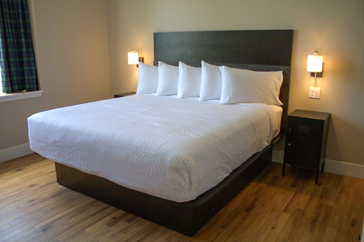 The spacious sleeping area features a king-sized bed with crisp white linens and multiple pillows. Wall-mounted bedside lamps provide soft lighting. Light-colored wood flooring complements the neutral wall tones, while a window dressed with plaid curtains allows natural light to enter.