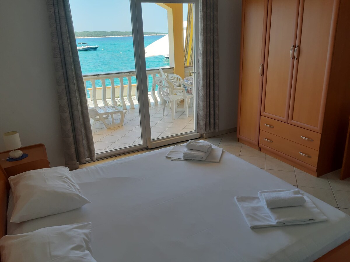 A bright sleeping area features a spacious bed with neatly arranged towels. Sliding glass doors open to a balcony with views of the turquoise sea and boats, allowing natural light to fill the room. Warm wood cabinetry offers storage space.