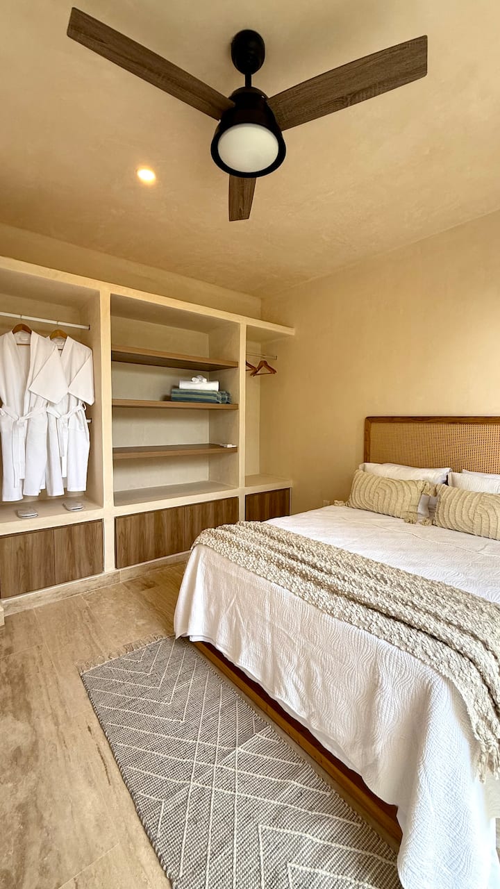 El dormitorio cuenta con una acogedora cama tamaño queen y sábanas suaves. La luz natural entra por las grandes ventanas, destacando la decoración minimalista pero elegante de la habitación. Con iluminación ambiental y toques serenos, es el espacio perfecto para descansar y recargar energías.

