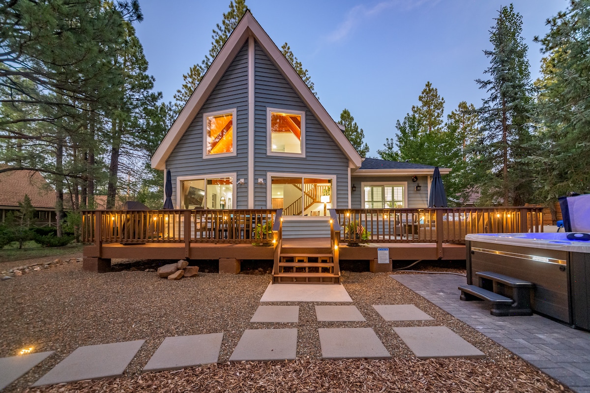 The exterior of The Cottage showcases a modern design with large windows and a spacious deck. Warm lighting highlights the architecture, surrounded by trees and gravel paths. A hot tub is visible nearby, inviting relaxation under the evening sky.