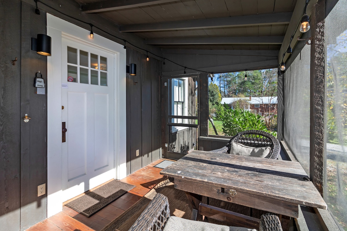 A shaded porch area is showcased, featuring a rustic wooden table surrounded by comfortable wicker seating. Decorative string lights add subtle illumination, while a door leads inside the cottage. A view of greenery is visible through the screened-in sections, enhancing a sense of privacy.