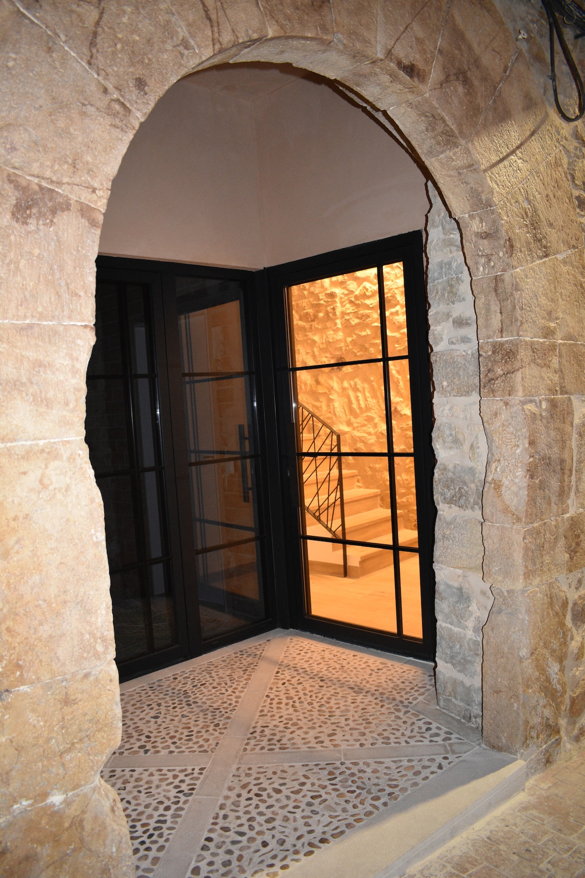 An inviting entrance features a large black-framed double door, providing a view of an illuminated staircase beyond. The stone archway complements the textured walls, while the floor is adorned with natural stone tiles arranged in a classic pattern.