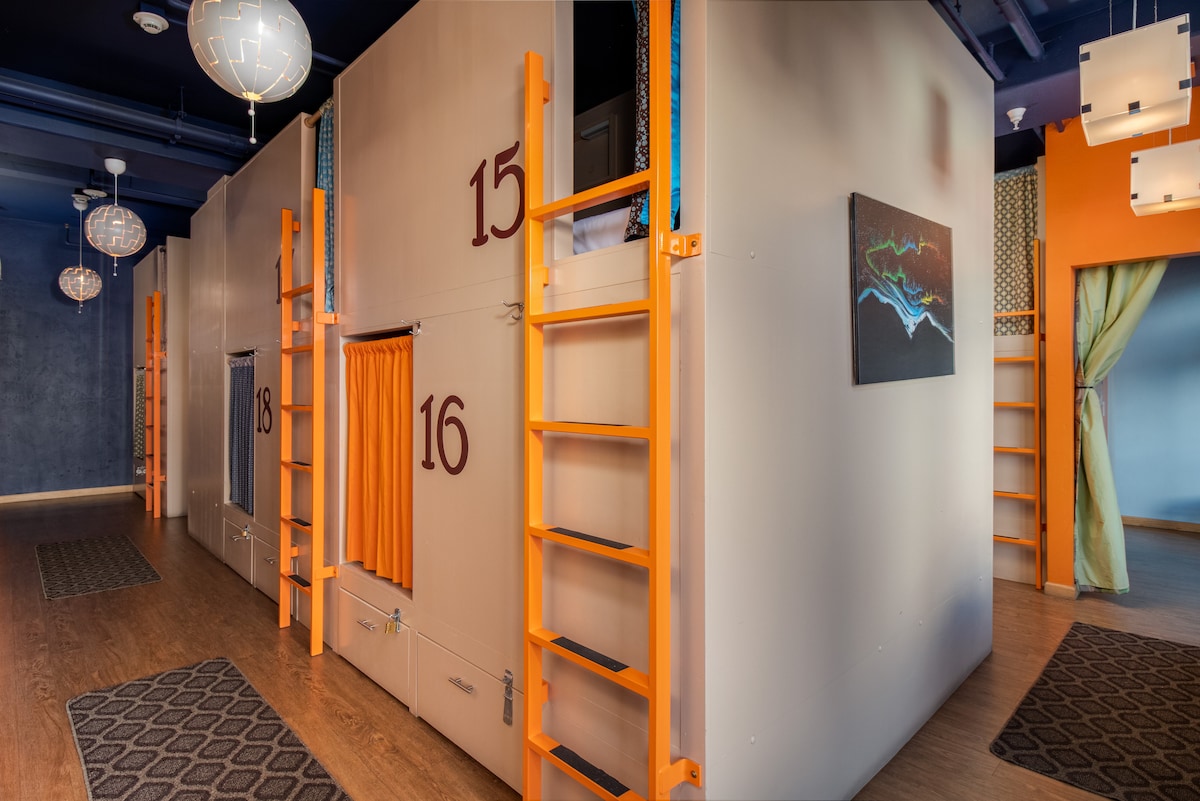 A shared living space is presented with custom wood-crafted bunk pods, each identified by numbers. Ladders provide access to upper bunks. Ambient lighting is provided by hanging fixtures. A colorful piece of wall art is displayed prominently, enhancing the room's modern design.