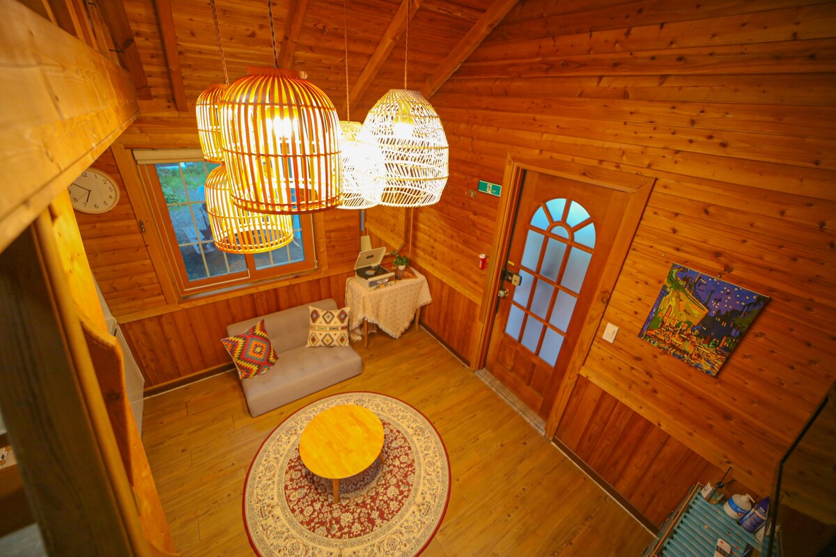A wooden interior is featured, highlighting a cozy seating area with a gray sofa and a round table at the center. Unique pendant lights hang from the ceiling, illuminating the space. A colorful painting adorns the wall, adding character to the welcoming atmosphere.