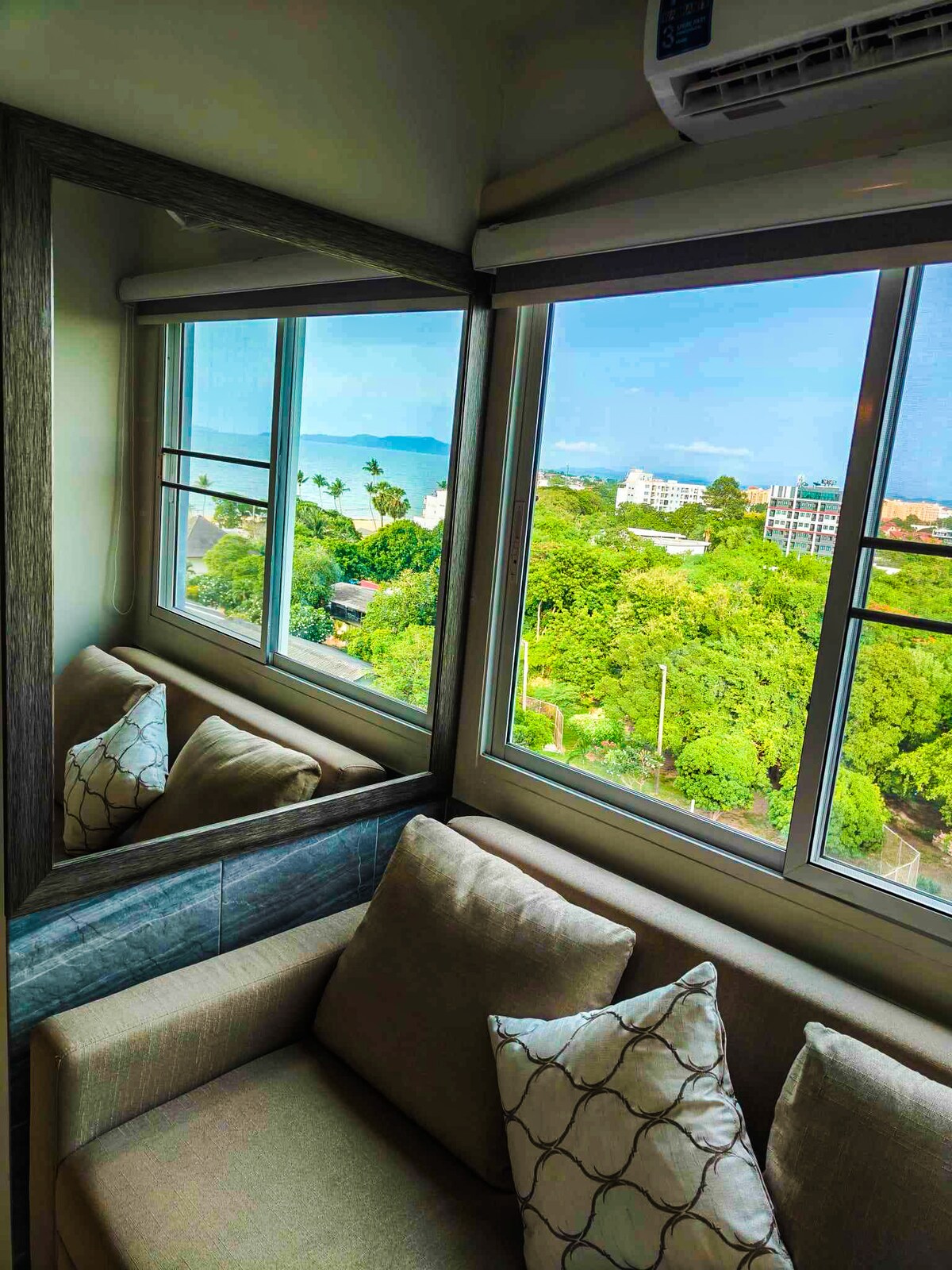 A corner seating area is visible, offering views through large windows adorned with a subtle decorative pillow on a light-colored sofa. The reflection of greenery and ocean can be seen in the glass, enhancing the sense of tranquility.