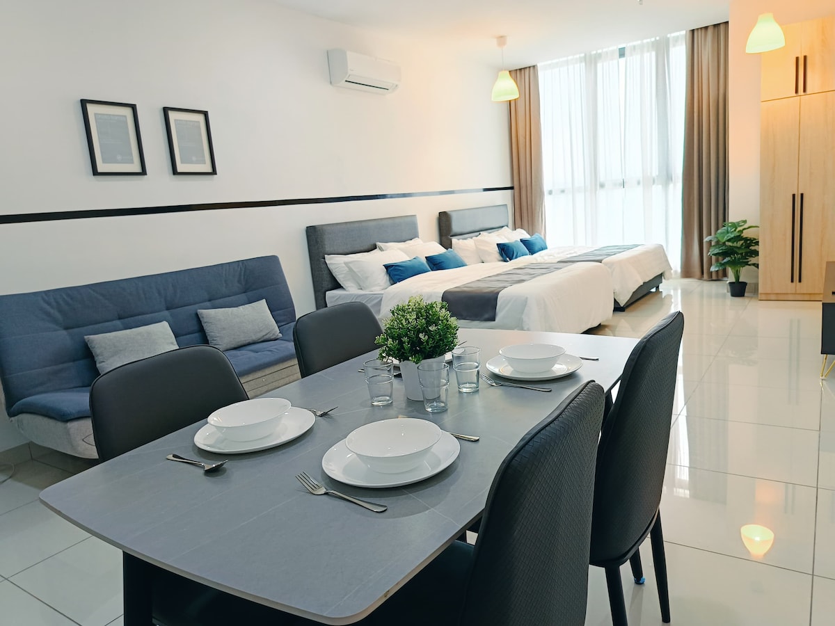 A spacious living area features a dining table set for four, accompanied by a cozy sofa and two queen-size beds. Large windows allow natural light to fill the room, with curtains adding a touch of warmth. Greenery is placed in a pot, enhancing the decor.