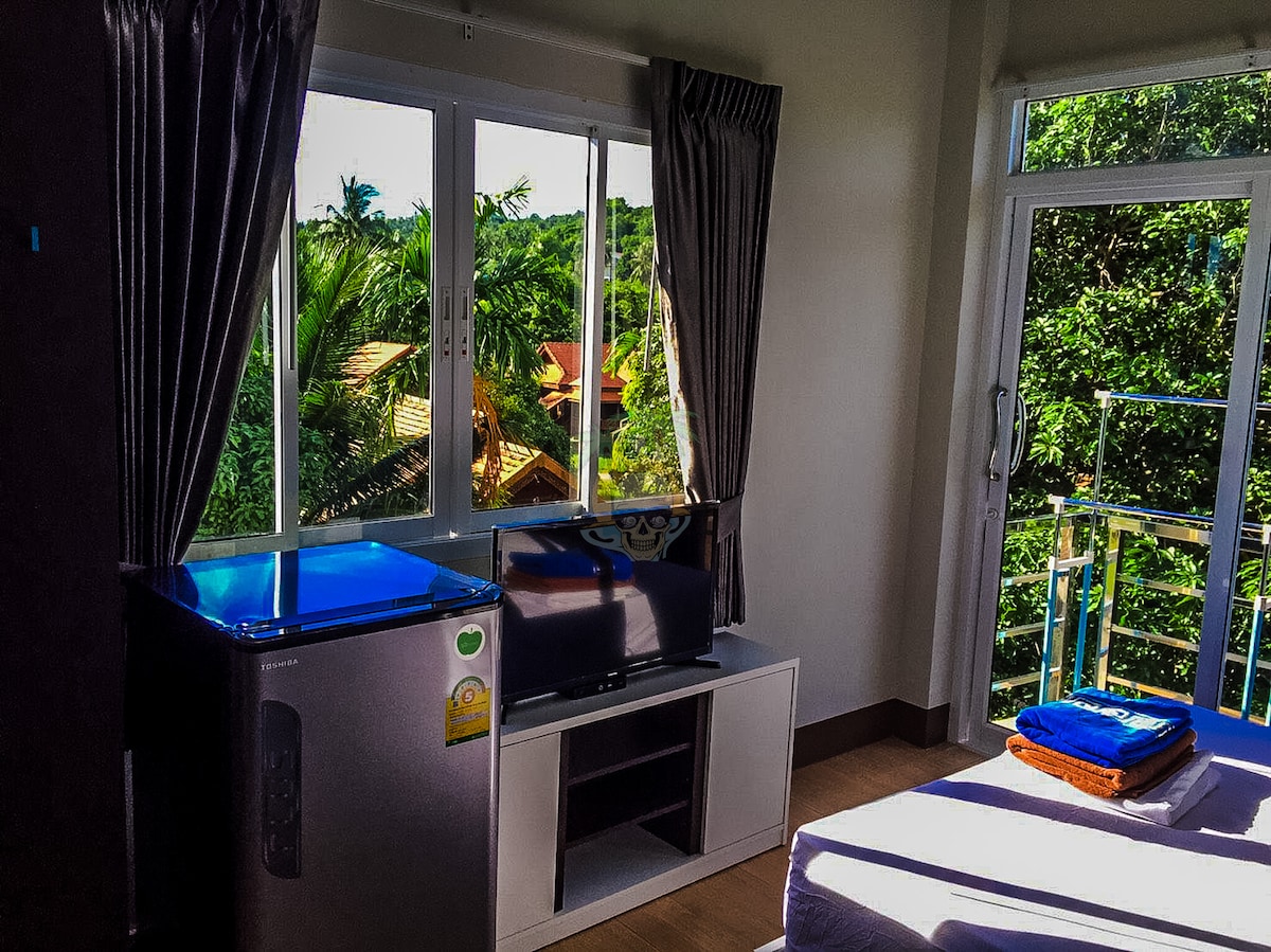 Natural light floods the room through large windows, revealing lush greenery outside. A compact refrigerator is positioned next to a flat-screen TV on a media console, with a neatly arranged stack of towels on a bed covered in crisp linens.