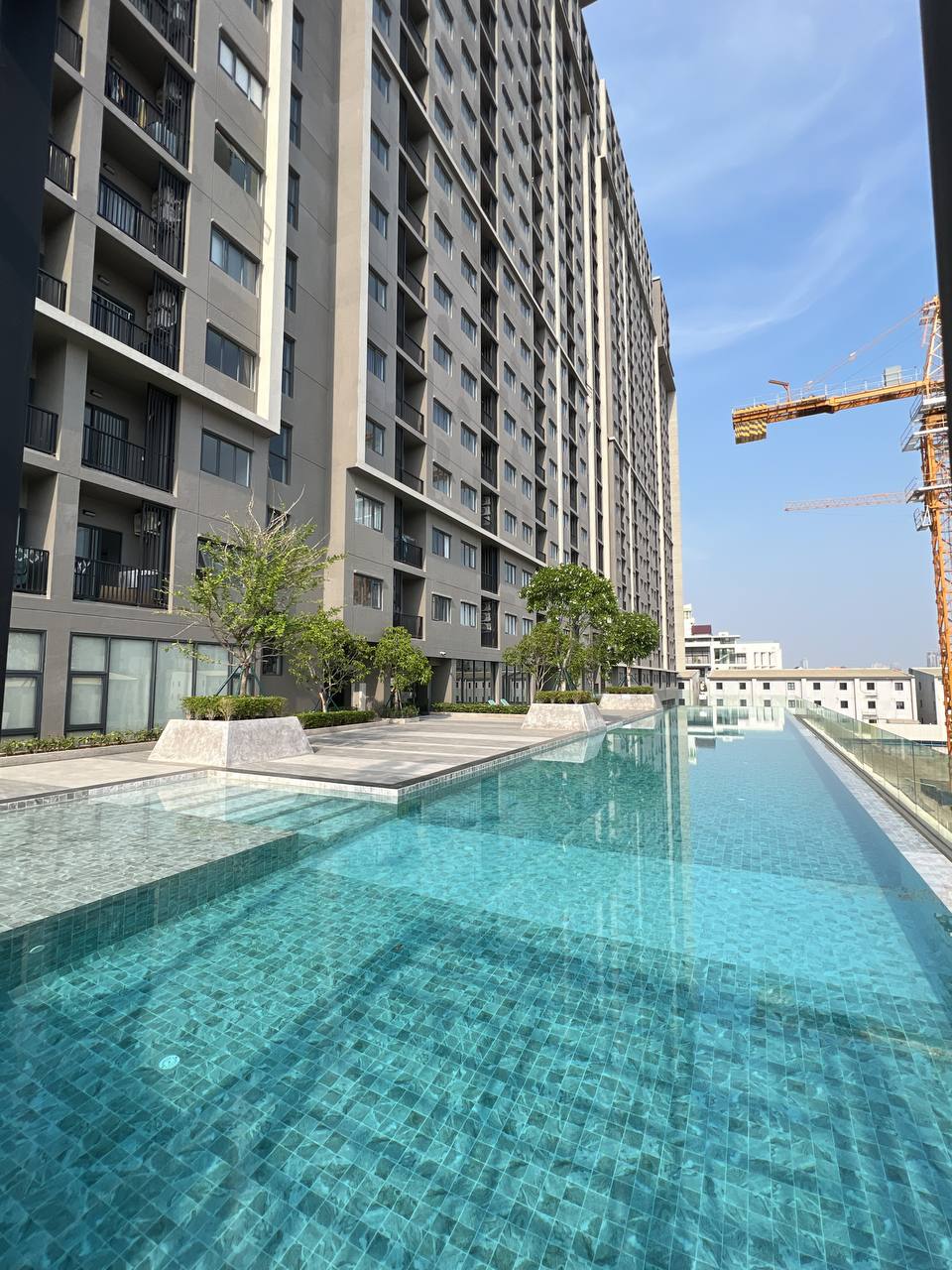 An outdoor pool is visible with clear blue water, reflecting the surrounding buildings. The adjacent architecture showcases modern design, featuring an expansive façade and balconies. A landscaped area with greenery borders the pool, contributing to a sense of openness.