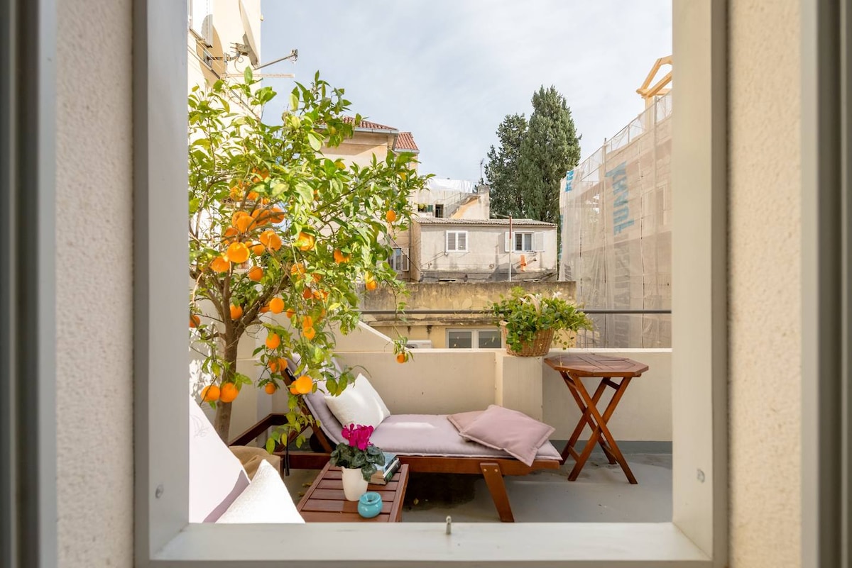 A private terrace is visible, featuring a wooden table and a reclining lounge chair adorned with cushions. An orange tree is seen bearing fruit, complemented by a potted plant with pink flowers on the table. A peaceful view of neighboring buildings is also present.