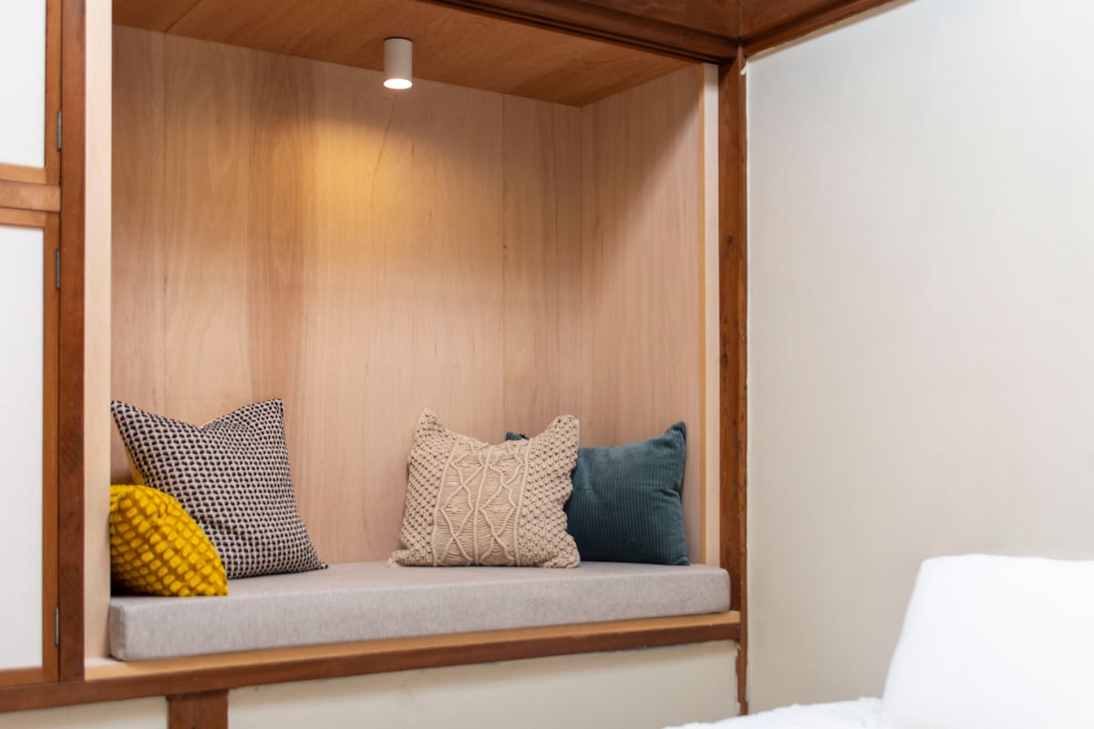 A cozy nook is presented with a cushioned bench, adorned with patterned pillows in neutral and green tones. A soft, warm light is mounted above, creating an inviting space for relaxation or reading.